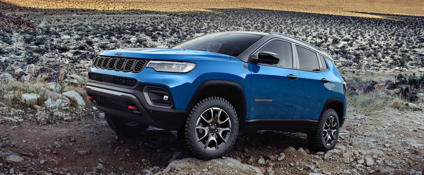 A blue 2025 Jeep Compass Trailhawk with a black hood insert, crawling over large and small rocks and stones off-road.