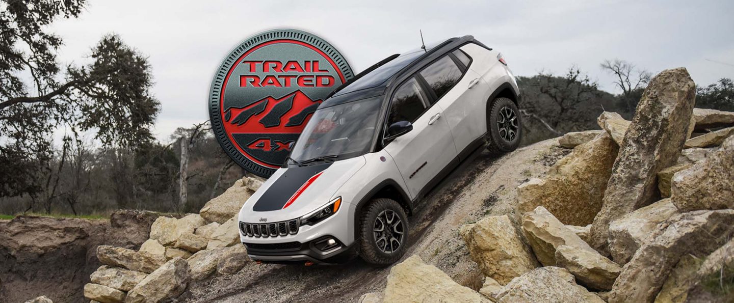 A white 2025 Jeep Compass Trailhawk with a black hood insert and black roof, descending a very steep hill off-road. The Trail Rated 4x4 badge.