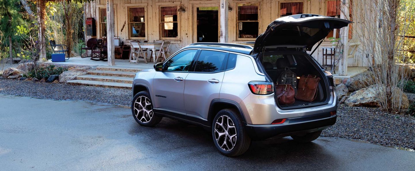 A silver 2025 Jeep Compass Limited parked at an old country store. Its liftgate is open revealing several pieces of luggage in the cargo area.