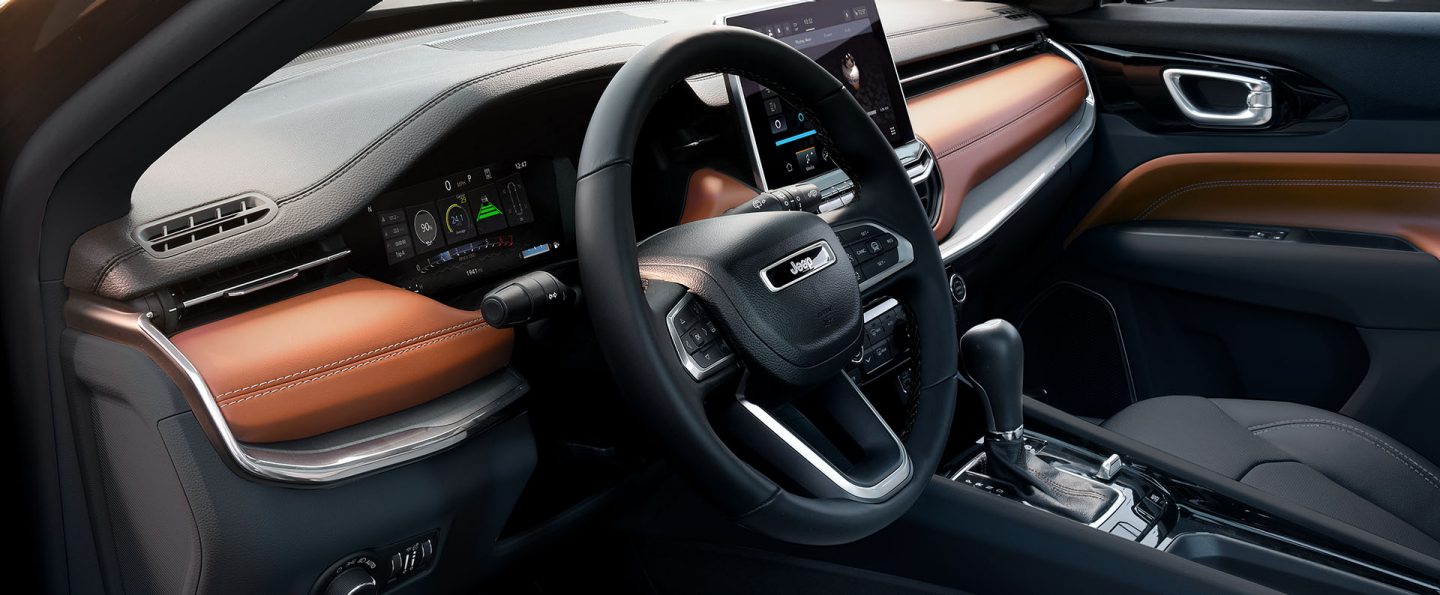 A raised angle of the driver seat area in the 2025 Jeep Compass Limited, concentrating on the steering wheel, Digital Cluster Display, Uconnect touchscreen and center console.
