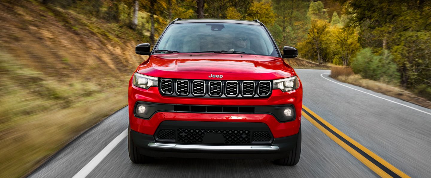 A head-on angle of a red 2025 Jeep Compass Limited traveling on a winding highway in the country. The background is blurred to indicate the vehicle is in motion.