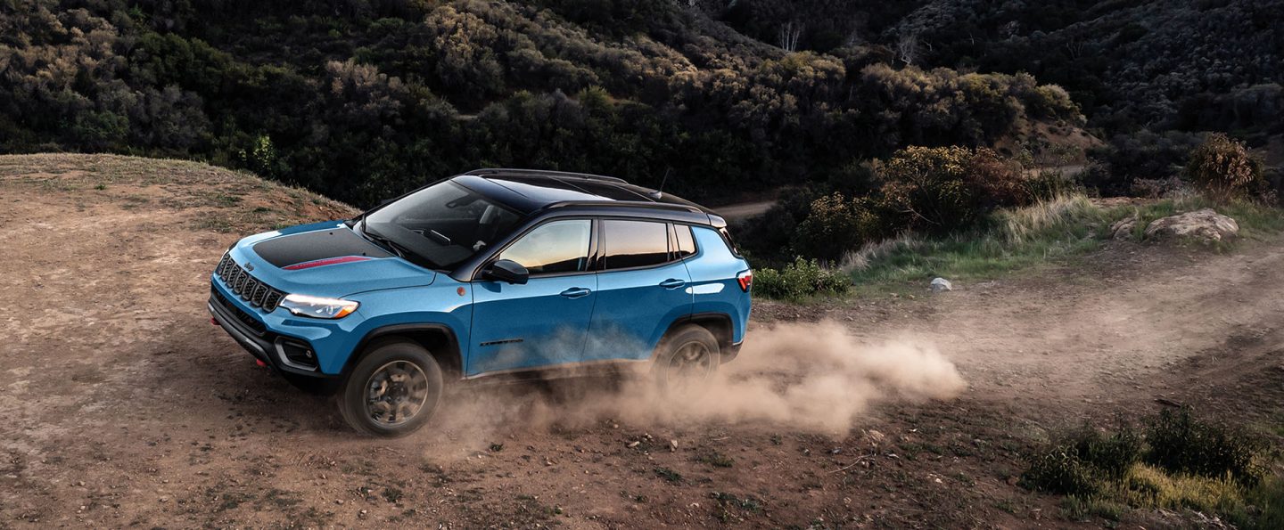 A blue 2025 Jeep Compass Trailhawk with a black hood insert, ascending a hill on a trail off-road, with the tires kicking up dust.