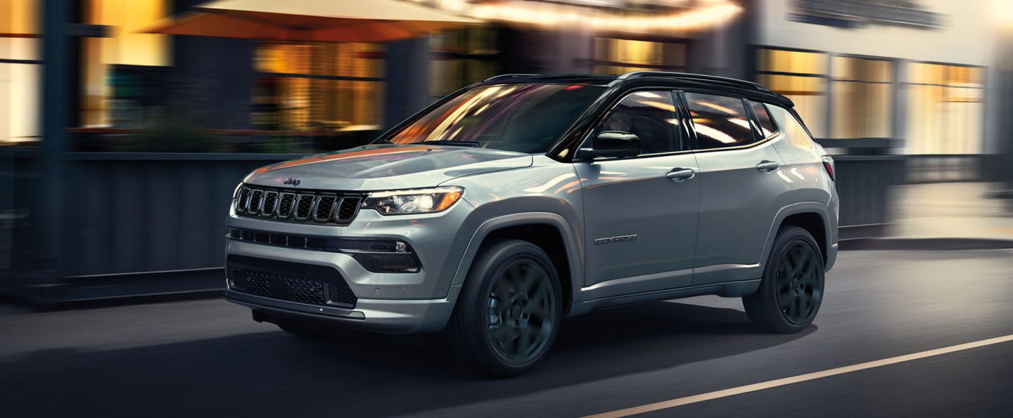 A silver 2025 Jeep Compass High Altitude traveling on a city street at night, with the background blurred to indicate the vehicle is in motion.