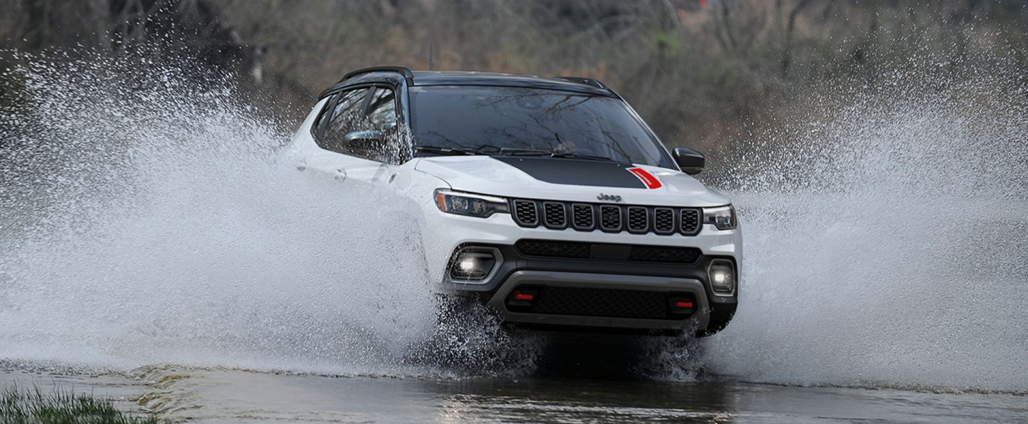 A white 2025 Jeep Compass Trailhawk with a black hood insert and black roof, traveling through a stream with water splashing up the sides of the doors.