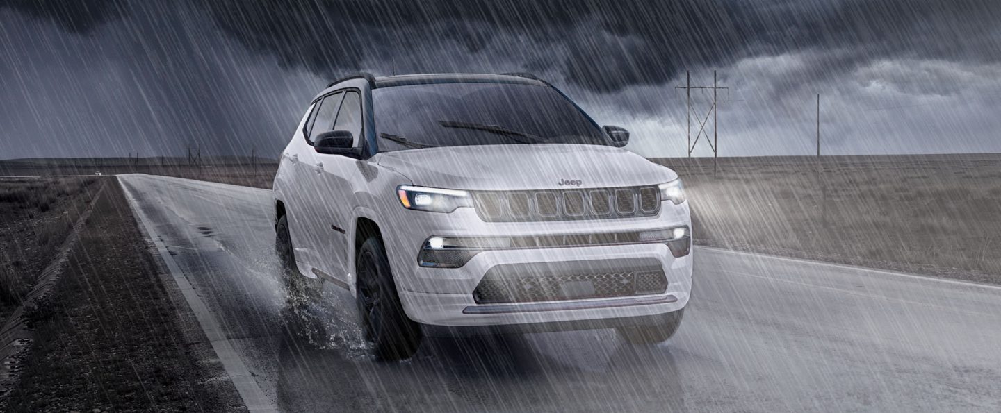 A white 2025 Jeep Compass High Altitude traveling through a dark, heavy rainstorm on a highway between two open fields.