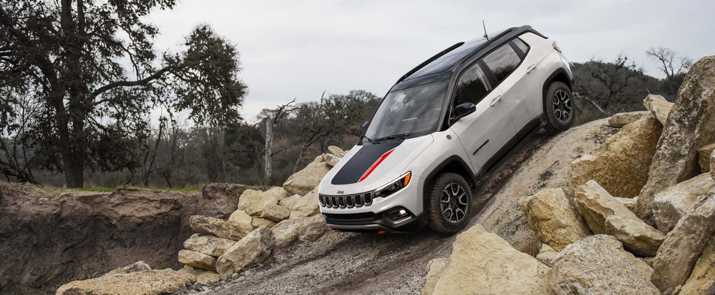 A white 2025 Jeep Compass Trailhawk with a black hood insert and black roof, descending a very steep hill off-road.