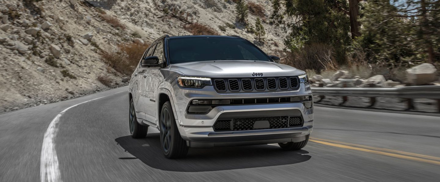 A front angle of a silver 2025 Jeep Compass High Altitude traveling on a winding mountain highway. The background is blurred to indicate the vehicle is in motion.