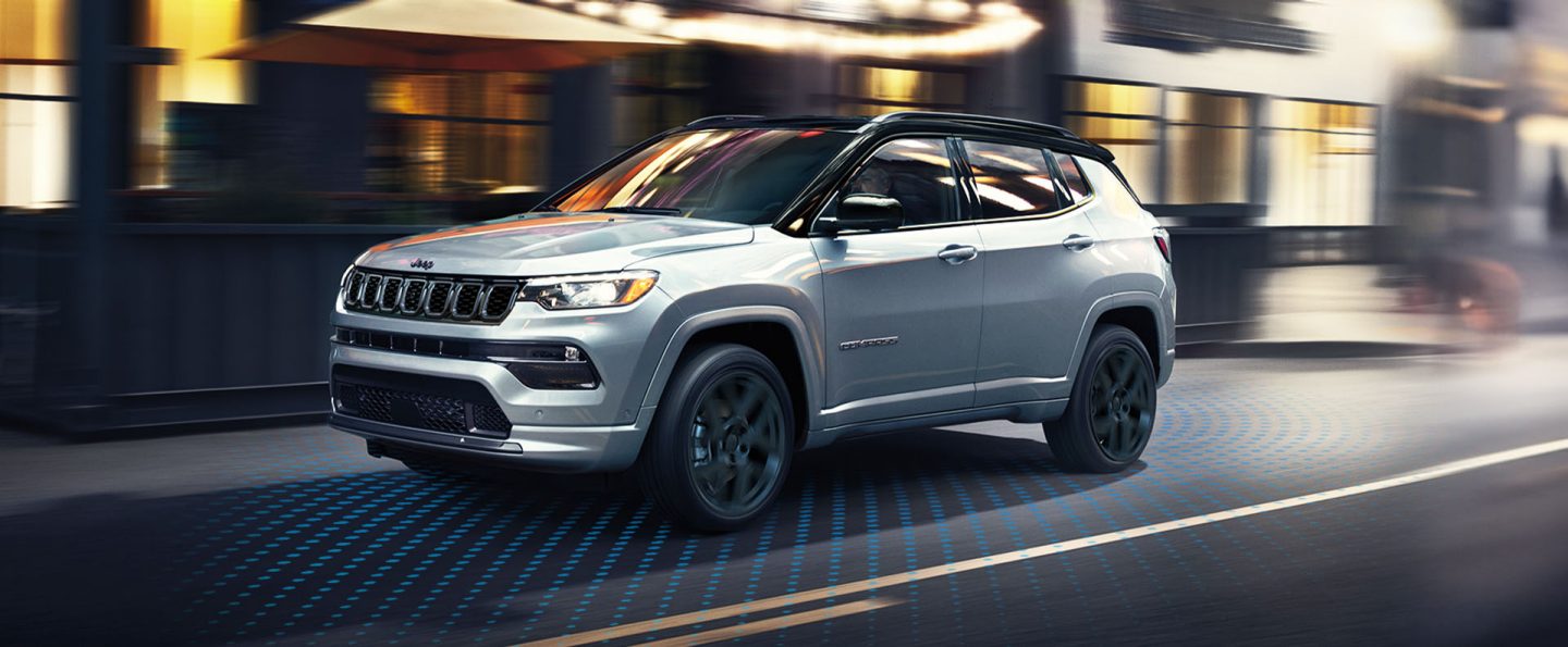 A silver 2025 Jeep Compass High Altitude traveling on a city street at night, with illustrated blue dotted lines emanating from all sides of the vehicle, to indicate sensors monitoring the surrounding area.