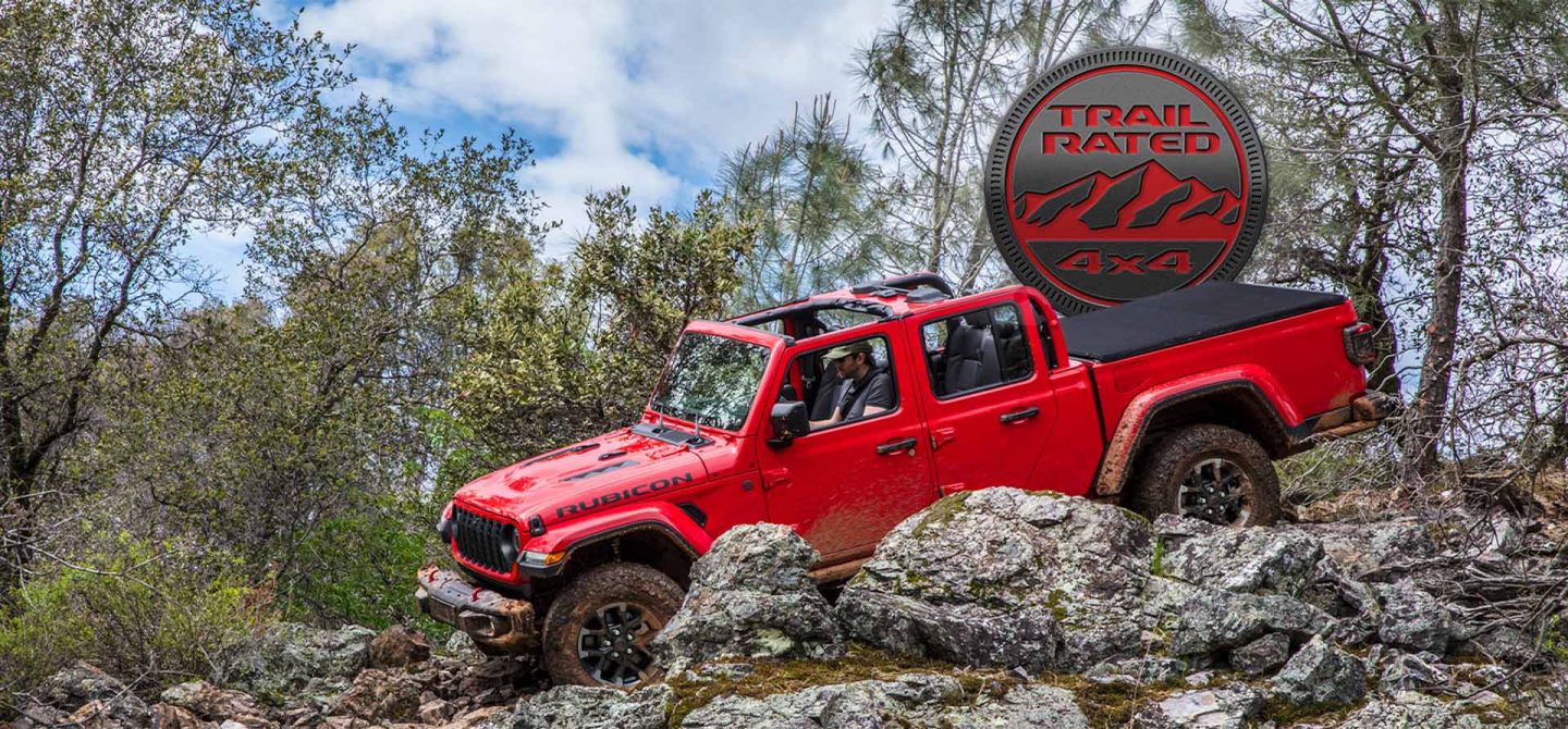 A red 2025 Jeep Gladiator Rubicon X with its roof removed, crawling over a series of boulders and outcroppings off-road. The Trail Rated 4x4 badge.