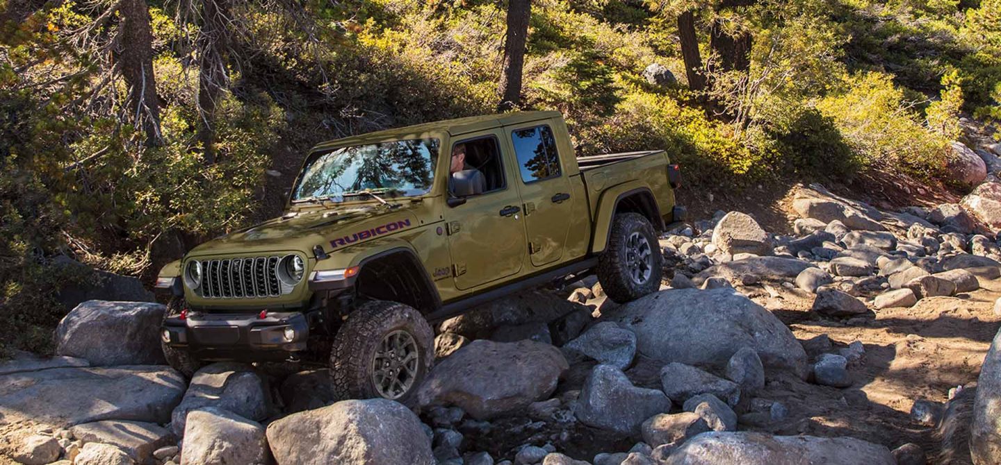 An olive green 2025 Jeep Gladiator Rubicon X crawling over a series of boulders in a forest off-road.