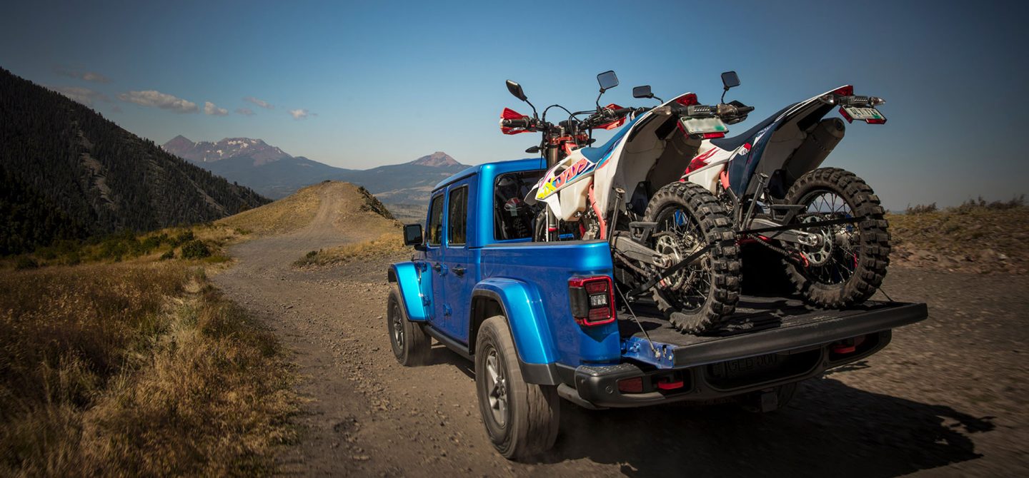 The rear of a blue 2025 Jeep Gladiator Mojave X being driven on a trail off-road, with its tailgate open and two ATVs tied down in its pickup bed.