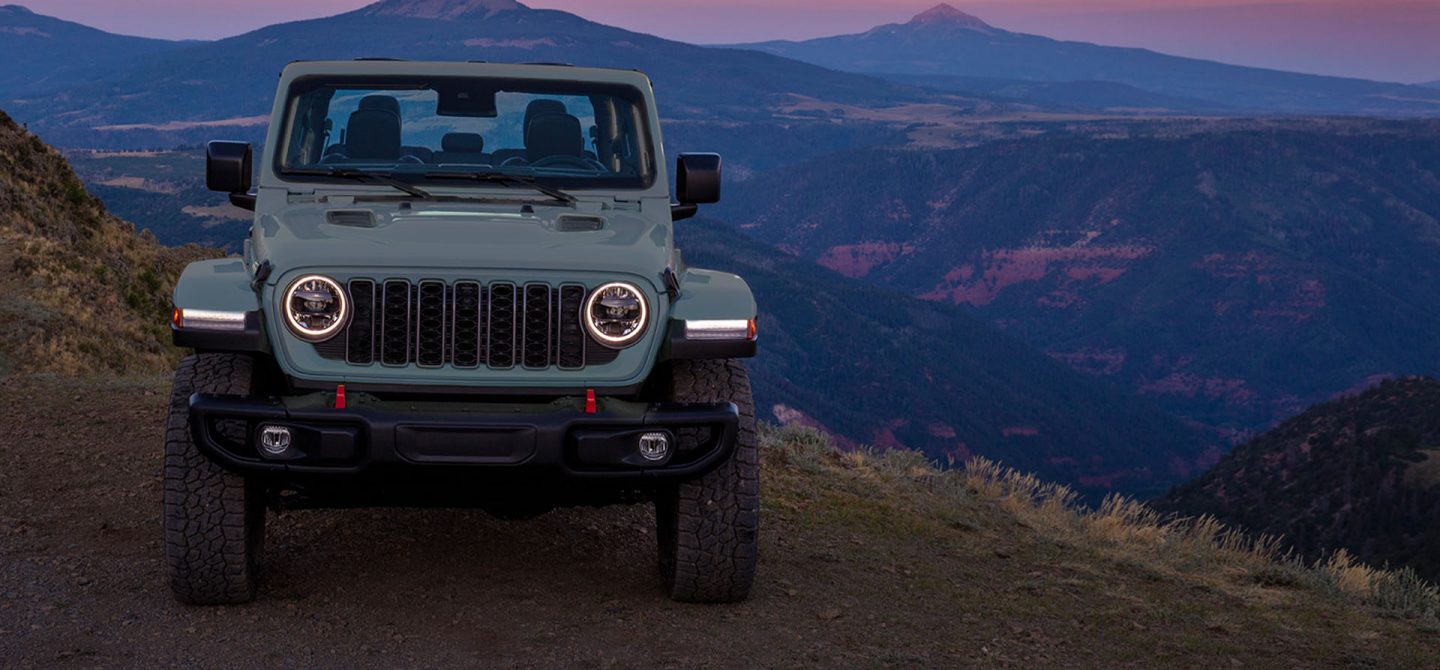 The front of a gray 2025 Jeep Gladiator Rubicon X parked on the edge of a cliff with mountains in the distance.