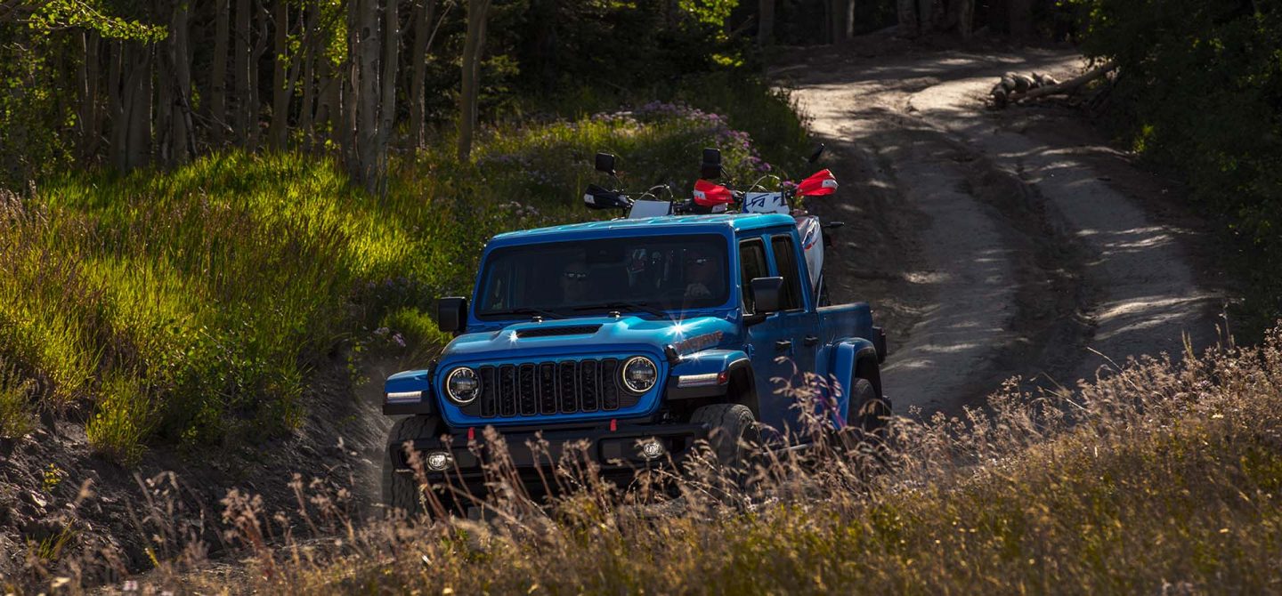 A blue 2025 Jeep Gladiator Mojave X being driven on a winding trail with two ATVs in its pickup bed.