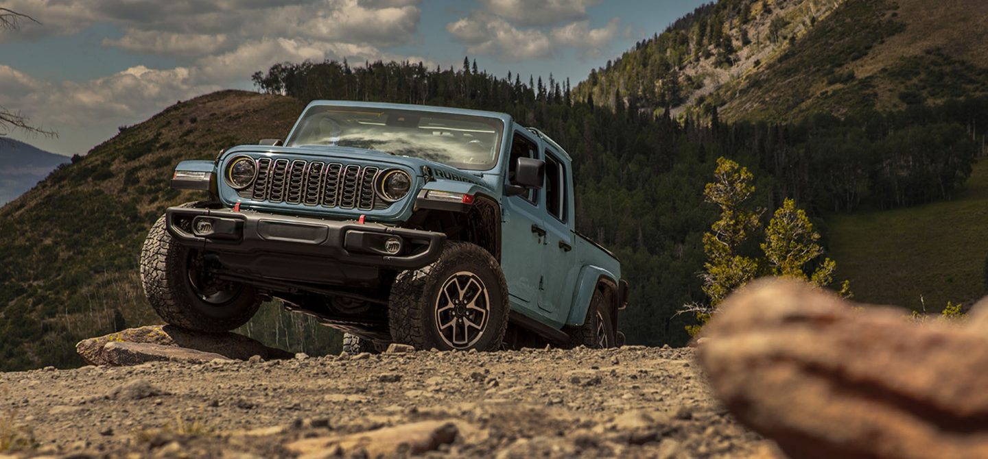 A red 2025 Jeep Gladiator Rubicon X ascending a rugged, rocky trail off-road, with an olive green 2025 Jeep Gladiator Rubicon X following behind.