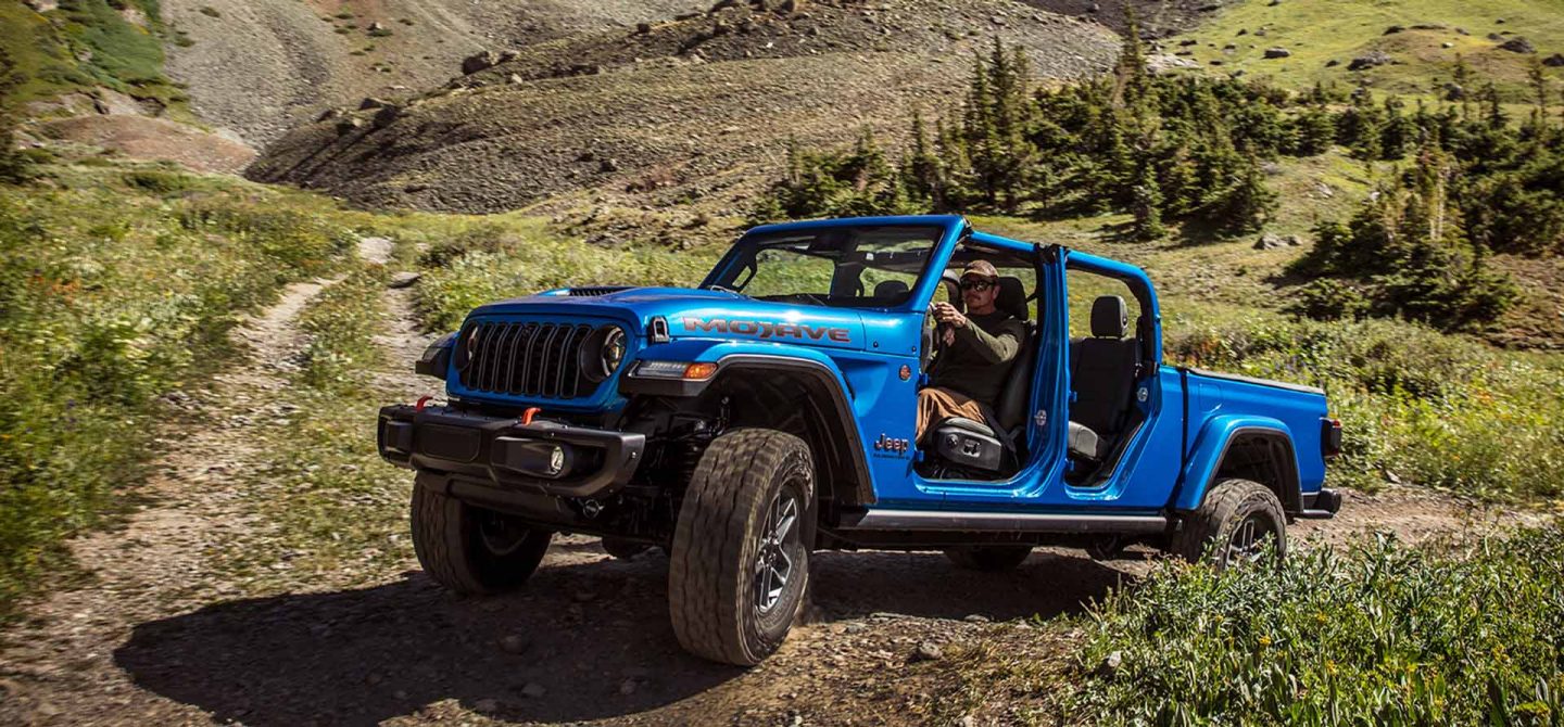 A blue 2025 Jeep Gladiator Mojave X with its doors and roof removed, ascending a trail in a hilly area off-road.