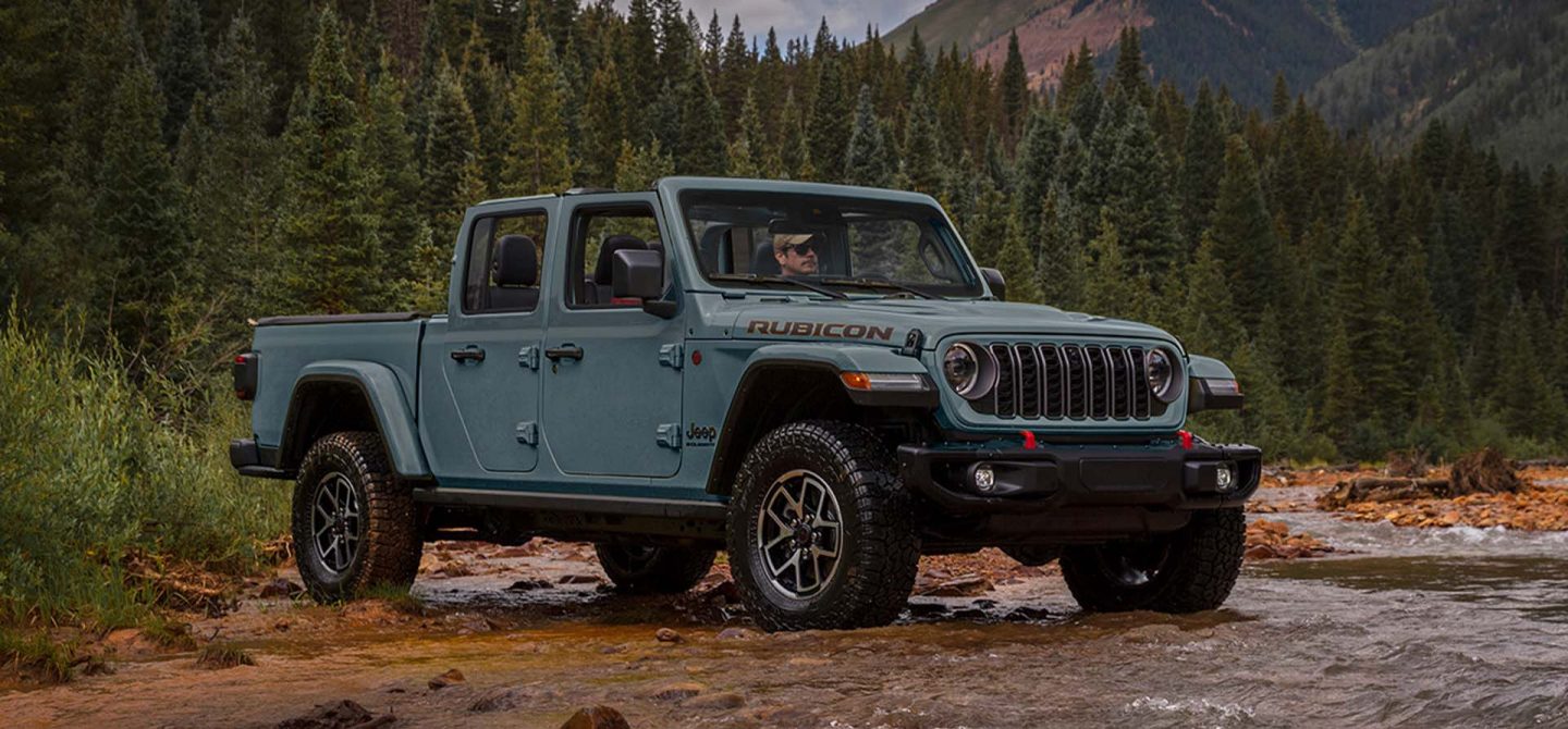 A blue 2025 Jeep Gladiator Mojave X with its doors and roof removed, ascending a trail in a hilly area off-road.