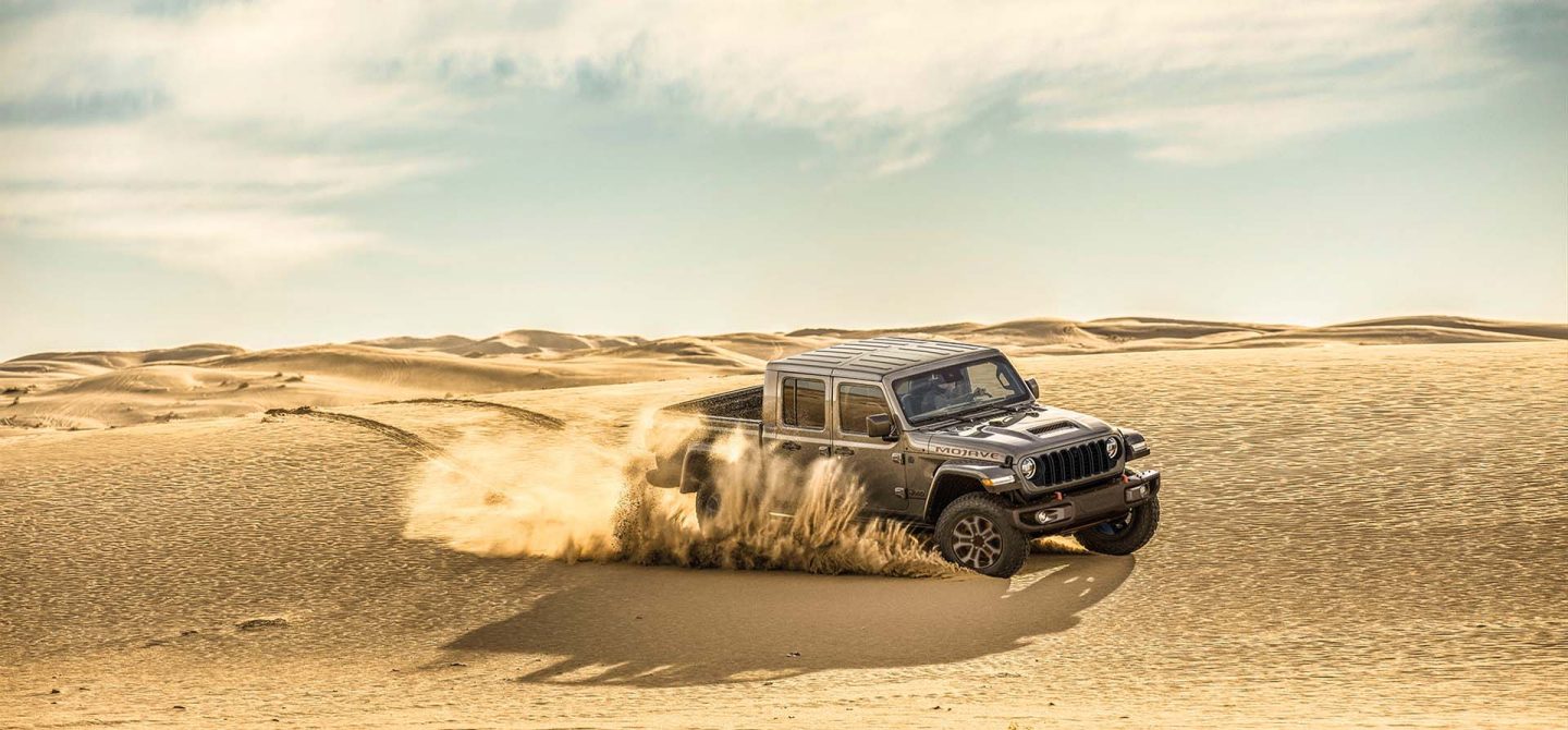 A gray 2025 Jeep Gladiator Mojave X kicking up sand as it is driven across a desert plain.