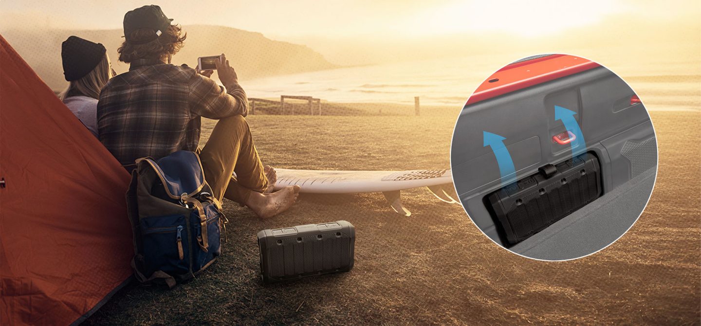 A couple sitting on a beach beside a tent, a removable wireless speaker next to them on the sand. An inset image displays where the speaker fits into the side of the 2025 Jeep Gladiator truck bed.