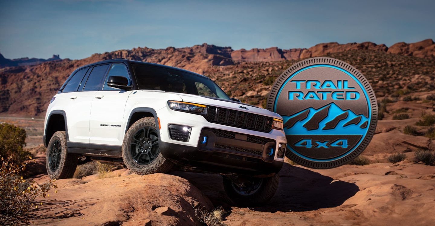 A white 2025 Jeep Grand Cherokee Trailhawk 4xe with black hood insert, rock crawling on uneven surfaces, over large boulders in the desert. The Trail Rated 4x4 badge.