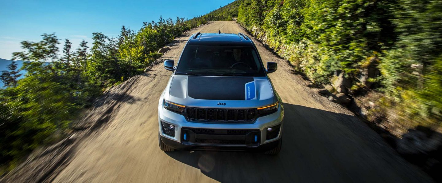 A head-on angle of a silver 2025 Jeep Grand Cherokee Trailhawk 4xe with black hood insert, traveling down a dirt road in the mountains, near a body of water.