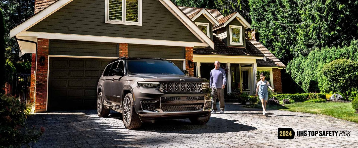 A gray 2025 Jeep Grand Cherokee Summit Reserve parked in the driveway of a residential home with a father and daughter nearby. 2024 IIHS Top Safety Pick.