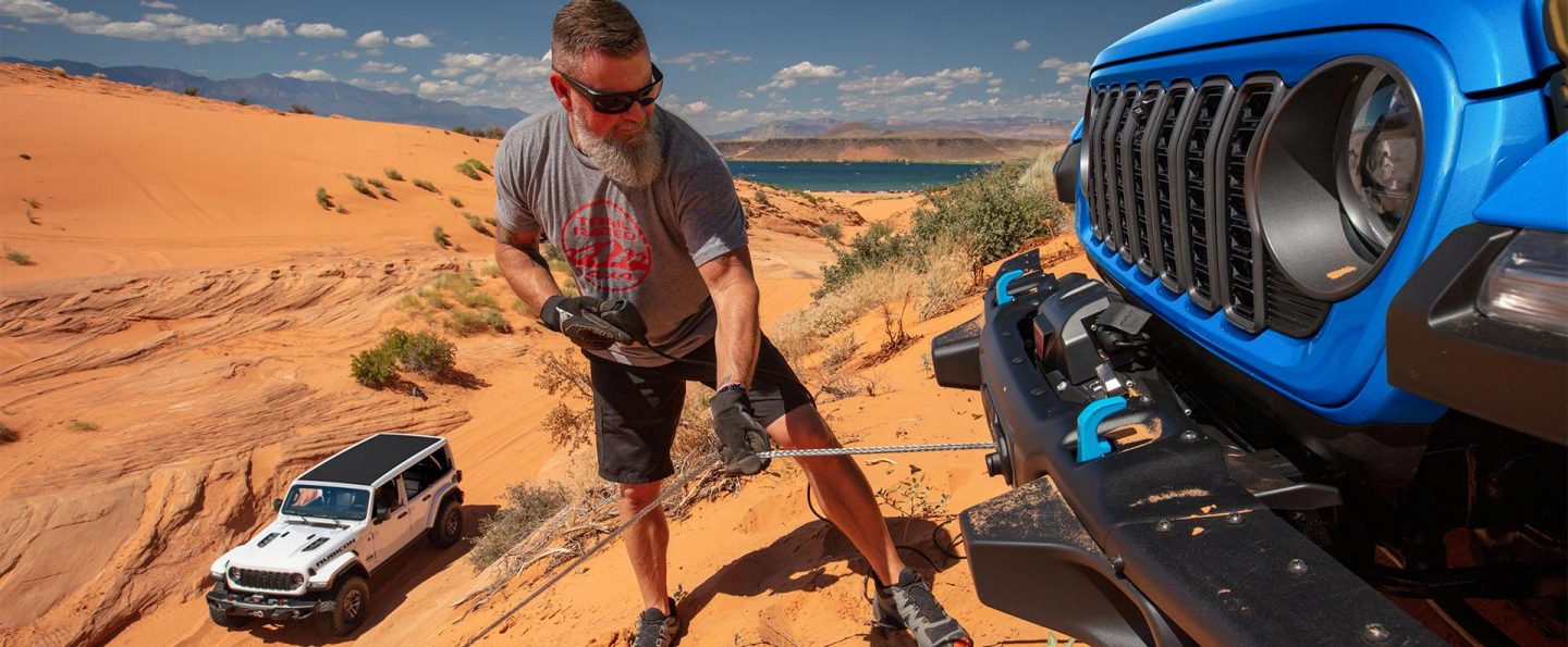 A man pulling the winch that's connected to the steel front bumper on a blue 2025 Jeep Wrangler Rubicon X 4xe as it sits at the top of a sand dune. A white 2025 Jeep Wrangler Rubicon X with black roof is parked below.