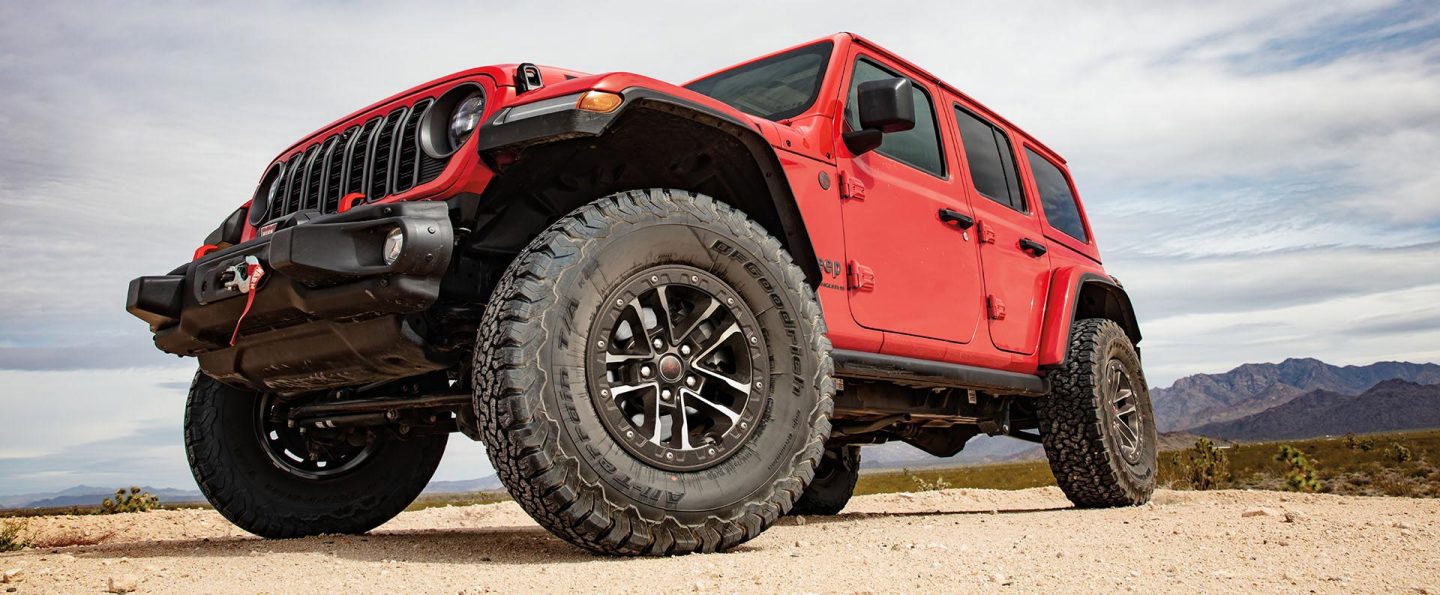 A low-lying front angle of a red 2025 Jeep Wrangler Rubicon X parked on sand, with mountains in the background.