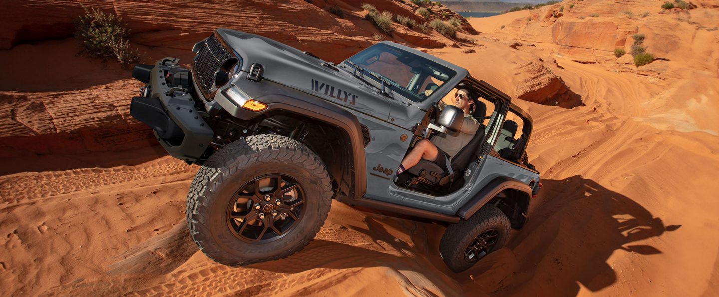 A gray 2025 Jeep Wrangler Willys with its doors and roof removed, crawling up and over a series of sand dunes in the desert.