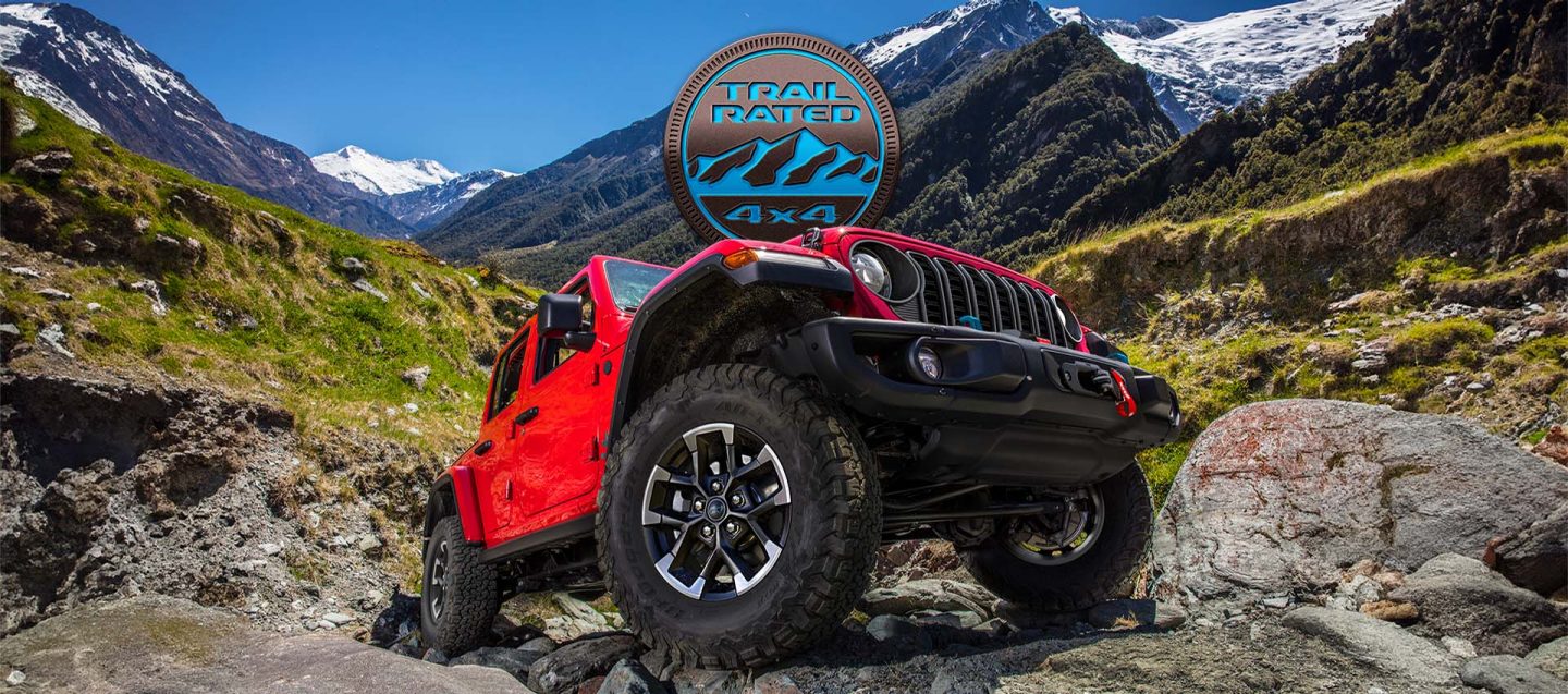 A low-lying angle of the wheels, tires and undercarriage of the 2025 Jeep Wrangler Rubicon X 4xe climbing a rocky trail off-road. The Trail Rated 4x4 badge.