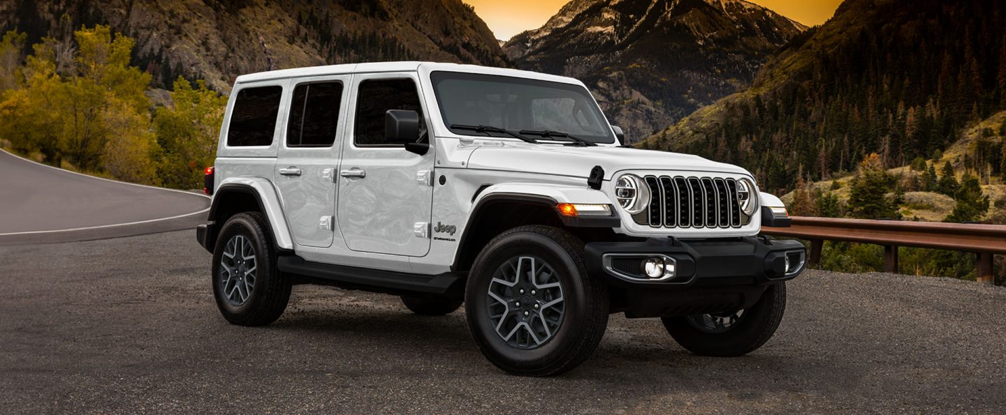 A passenger-side front angle of a white 2025 Jeep Wrangler Sahara parked on the shoulder of a highway in the mountains at sunset.