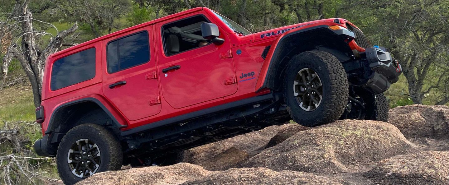 A passenger-side profile of a red 2025 Jeep Wrangler Rubicon X 4xe climbing a hill of sand-covered boulders.