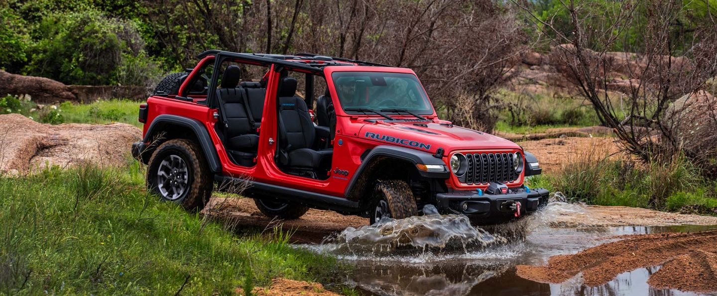 A red 2025 Jeep Wrangler Rubicon 4xe with its doors and roof removed, entering a stream off-road.