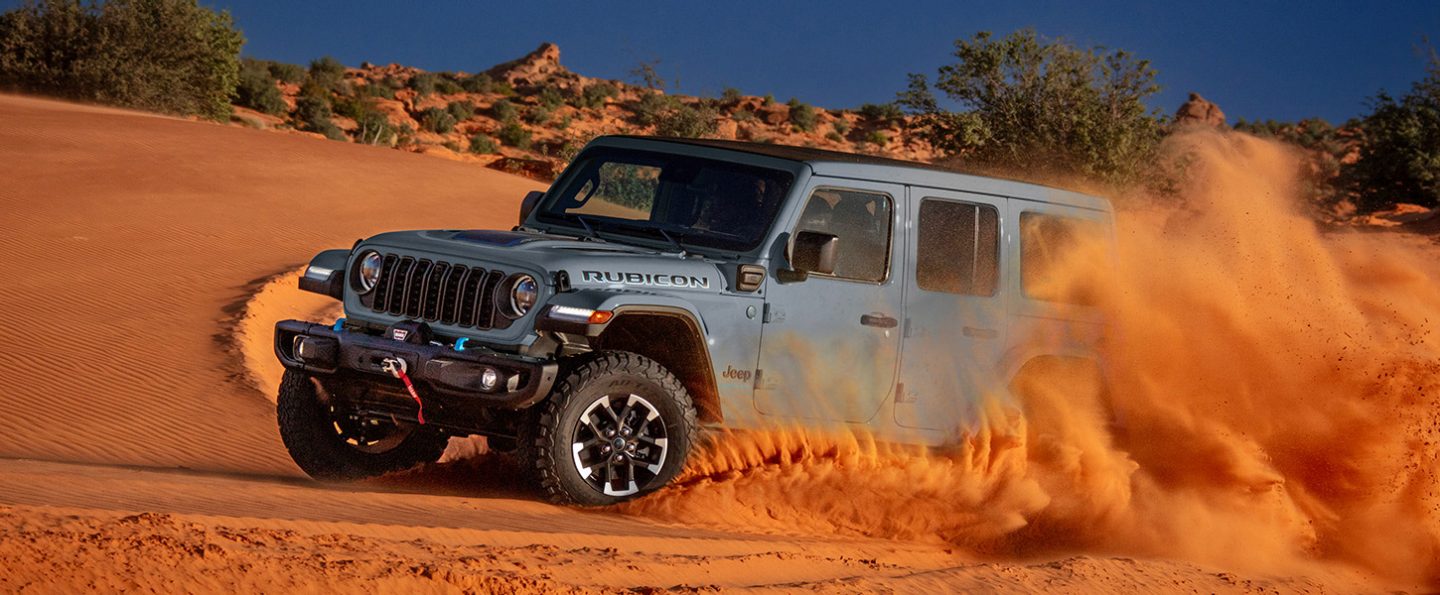 A gray 2025 Jeep Wrangler Rubicon X 4xe traveling across a dune in the desert, kicking up sand as it goes.