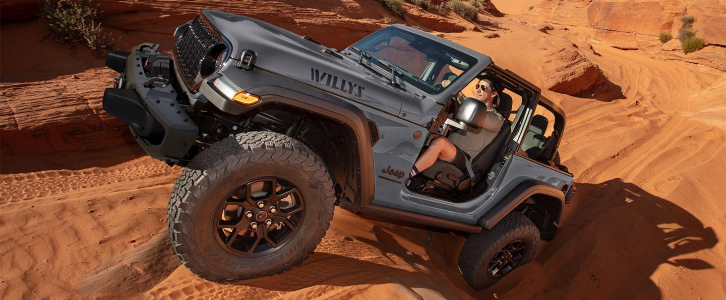 A gray 2025 Jeep Wrangler Willys with its doors and roof removed, crawling up and over a series of sand dunes in the desert.