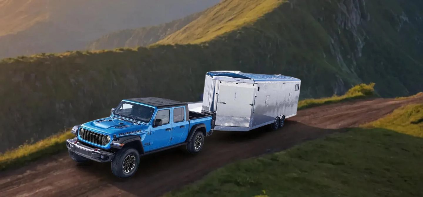 A blue 2025 Jeep Gladiator Rubicon being driven in a mountain as it tows an enclosed utility trailer.